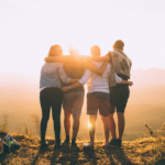 four person hands wrap around shoulders while looking at sunset — Foto por Helena Lopes na Unsplash